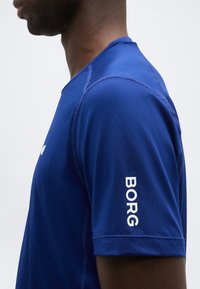 Πλάγια όψη ενός ατόμου που φοράει μπλε πουκάμισο με κοντά μανίκια και λευκή επιγραφή "BORG" στο μανίκι, με απλό φόντο.