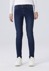 Los jeans ajustados de mezclilla azul oscura presentan un ligero brillo, costuras resistentes y un diseño de cinco bolsillos. Combinados con zapatillas blancas.