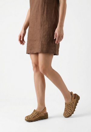 Kvinde iført en brun knælængde kjole og tan perforerede slingback sko med bølget sål, stående og gående mod en ensfarvet baggrund.