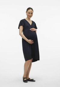 Marineblaue Umstandsbekleidung mit kurzen, schwingenden Ärmeln und einem V-Ausschnitt. Merkmale sind eine geraffte Taille und ein knie-langer Saum. Getragen mit schwarzen Ballerinas.