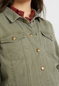Chaqueta verde oliva con botones de bronce, dos bolsillos en el pecho y un cuello, con un acabado texturizado y un cuello rosa con volantes debajo.