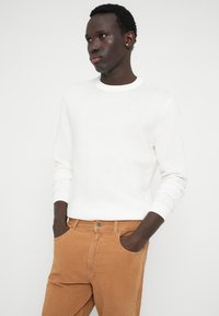 Bela, rebrasta pletenina s krojem okoli vratu, v kombinaciji s svetlom rjavimi kordnimi hlačami. Obleka ima dolge rokave in prilegajoč izgled.