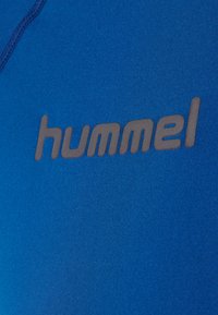 Blå t-shirt med en glat tekstur. Har et fremtrædende sort "hummel" logo trykt på fronten. Syninger er synlige langs kanterne.