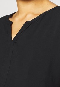 Haut noir avec un décolleté en V, en tissu doux et à coupe décontractée. Des coutures le long du décolleté et de la patte de boutonnage ajoutent une texture subtile.