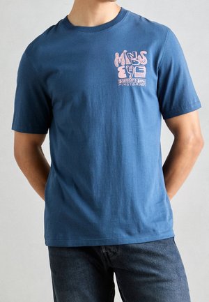 Mężczyzna w niebieskim T-shircie z różowym nadrukiem "Minds Eye Scotch & Soda Amsterdam" na lewej piersi, w ciemnych dżinsach.