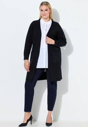 Femme portant un long cardigan noir sur une chemise blanche boutonnée, un pantalon ajusté foncé et des talons hauts noirs, debout devant un fond blanc.