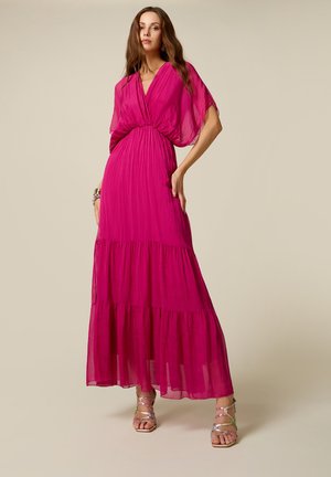 Kobieta stoi ubrana w jasnoróżową, wielowarstwową suknię maxi z krótkimi, zwiewnymi rękawami i metalicznymi sandałami na wysokim obcasie z paskami.