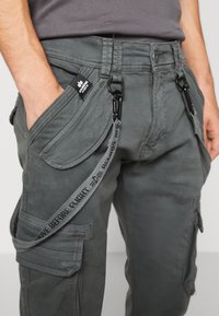 Pantalones cargo grises con múltiples bolsillos, tejido resistente, un lazo de sujeción con texto y un acabado texturizado. Presenta una etiqueta en la cintura.