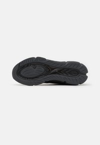 Semelle d'une chaussure de sport noire avec une bande de roulement texturée et des rainures incurvées pour l'adhérence, montrée sur un fond blanc uni.