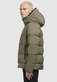 Olīvzaļa pūkainā jaka ar augstu apkakli un regulējamu kapuci; ar rāvējslēdzēja sānu kabatu un teksturētu, quilted dizainu.