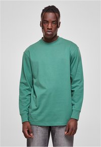 Camisa de algodón verde de manga larga con cuello redondo, corte relajado y puños acanalados, combinada con jeans gris desvanecido.