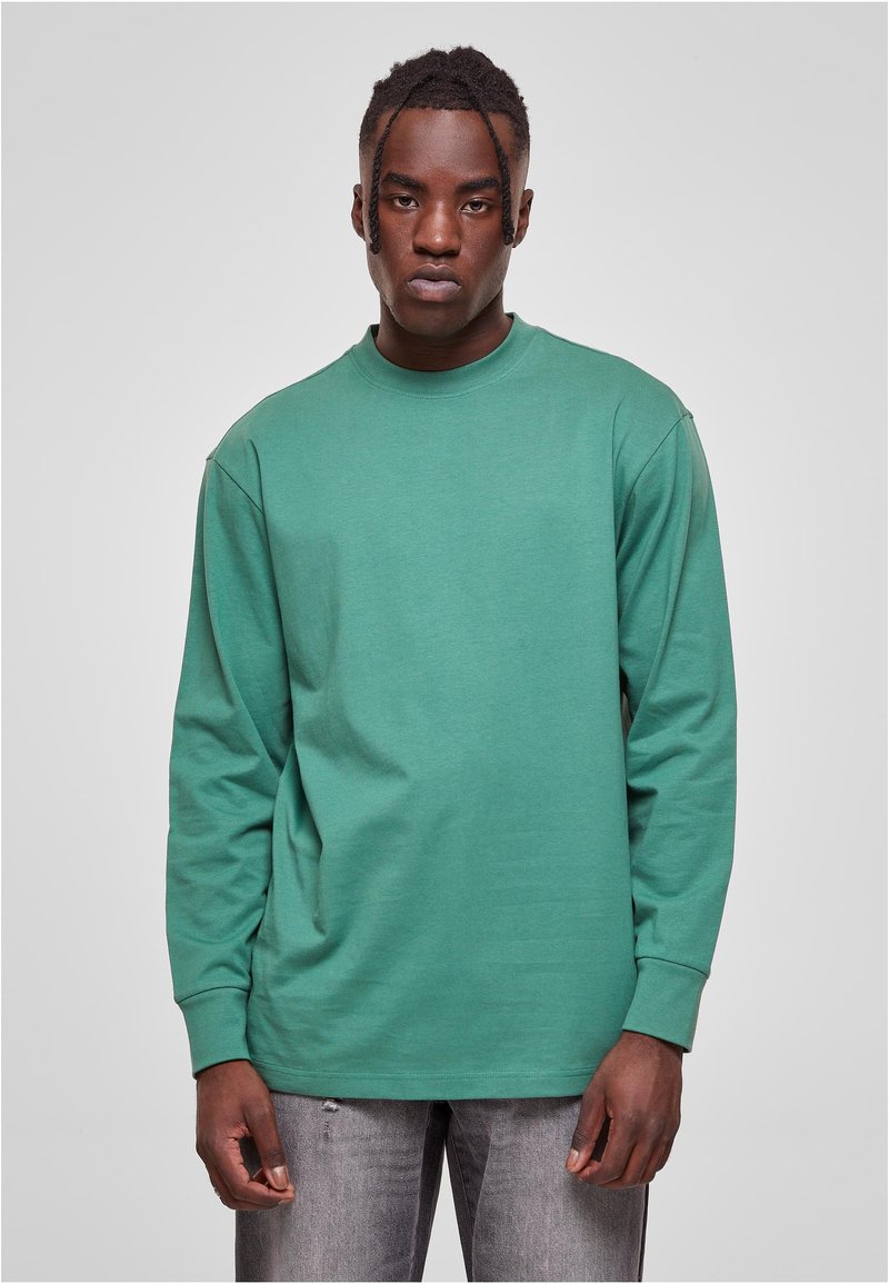 Camisa de algodón verde de manga larga con cuello redondo, corte relajado y puños acanalados, combinada con jeans gris desvanecido.