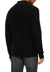 Zwarte gebreide trui met een geribbelde textuur, voorzien van een brede kraag en lange mouwen, gecombineerd met grijze denim jeans. Zichtbare stikseldetails.