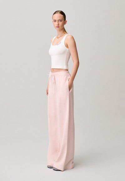 Mujer joven con camiseta sin mangas blanca ajustada, pantalones amplios de color rosa claro con cordón, zapatos blancos y negros, de pie frente a un fondo gris liso.