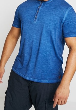 Camiseta henley de manga corta en azul, confeccionada con una suave tela texturizada. Presenta una tapeta de tres botones y un dobladillo redondeado, con variaciones de color sutiles.