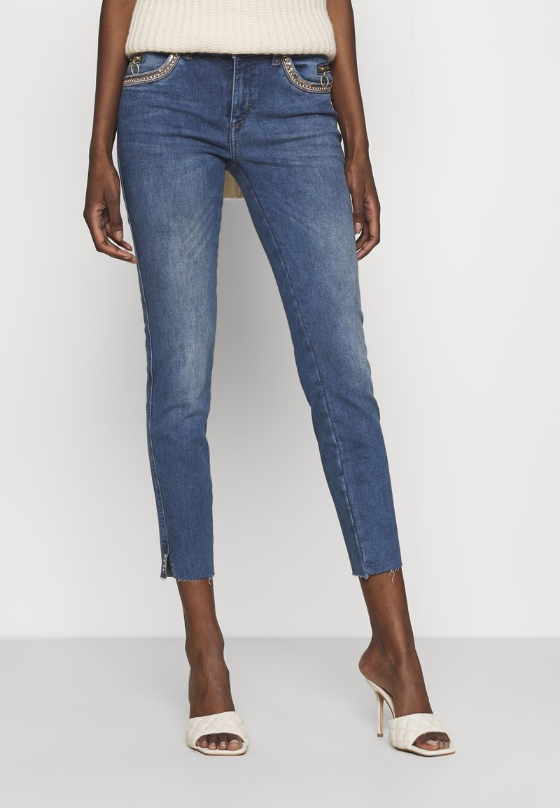 Jeans skinnies de mezclilla en azul oscuro, con acentos metálicos en los bolsillos, longitud recortada con dobladillo sin terminar y cremalleras en los tobillos.