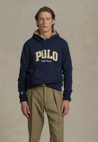 Sötétkék kapucnis pulóver "POLO" felirattal krém színben és Ralph Lauren márkázással, bézs kockás nadrággal, amelyen redők találhatók. Hétköznapi összeállítás.