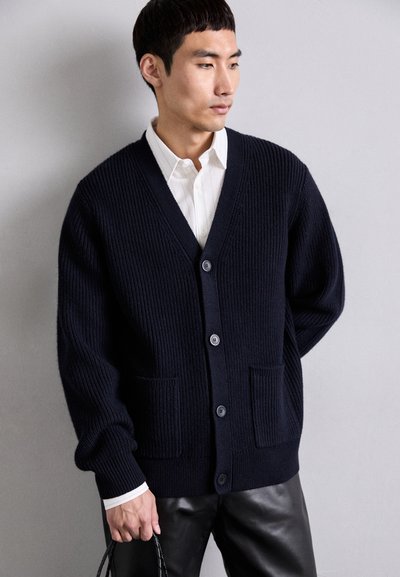 Tumesinine ribakardigan V-kaelusega, kuue nupuga ja kahe esimise taskuga, koos valge kraega särgi ja mustade nahkpükstega.