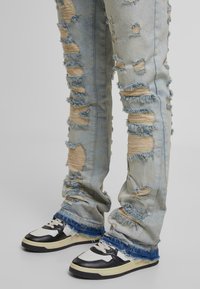 Šviesiai mėlyni nusidėvėję džinsai su suplėšytais ir nuskaldytais akcentais, derinami su juodai baltais sportiniai bateliai, turintys tekstūruotą pado ir raištelių modelį.