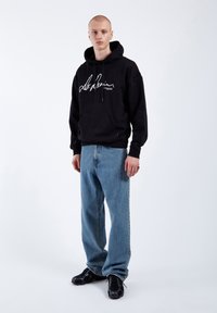 Black hooded sweatshirt with a white signature logo, paired with light blue straight-leg jeans and black sneakers. Plain background.