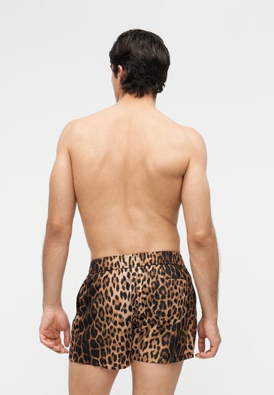 Oberkörperfreier Mann mit kurzen dunklen Haaren, der leopardgemusterte Badeshorts trägt und mit dem Rücken zur Kamera vor einem schlichten Hintergrund steht.