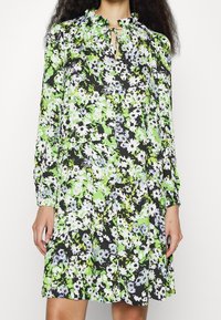 Long-sleeve dress in black, adorned with a floral pattern in green, white, and blue. Features a ruffled neckline and a keyhole opening.