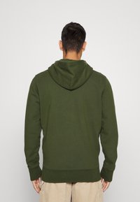 Homme portant un sweat à capuche vert foncé uni et un pantalon beige, debout de dos devant un fond clair uni.