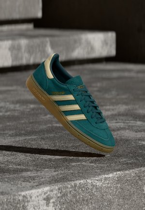 Petrolinvärinen Adidas mokkanahkainen tennari, jossa on kermaan vivahtavat raidat ja kumipohja, leijuu betonisella ulkoportaalla.