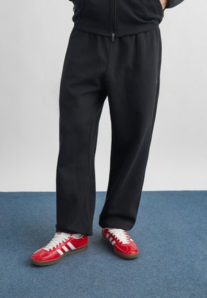 Osoba stojaca na modrom koberci s rukami v kapsách, oblečená v čiernych teplákoch a červených teniskách Adidas.