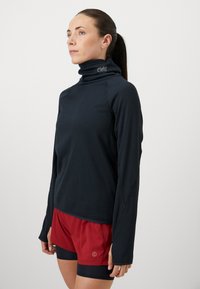 Sort langærmet skjorte med høj hals lavet af tekstureret stof, parret med røde shorts. Har logo på kraven og kontrasterende syninger.