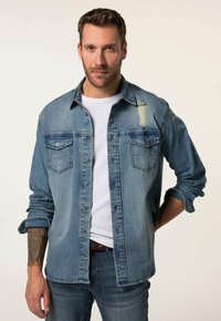 Camisa de mezclilla azul clara con cierres de botón, dos bolsillos en el pecho y un detalle desgastado en el hombro; llevada sobre una camiseta blanca.