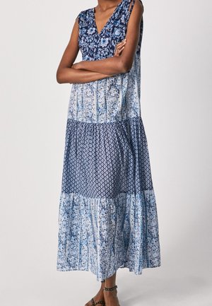 Mouwloze lange jurk met drie blauwe panelen met patroon, V-hals, gedragen door een persoon met gekruiste armen en zwarte sandalen met studs.