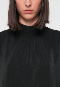 Donna con capelli castani lisci che indossa un blusa nera a collo alto, labbra chiuse, su uno sfondo chiaro e semplice.