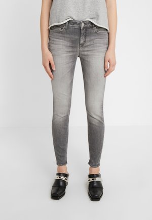Femme portant un jean skinny gris délavé, un top gris et des chaussures slip-on noires avec un détail chaîne argentée, debout sur fond blanc.
