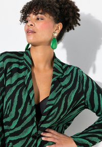 Blazer a righe zebrate verdi e nere con colletto a punta e chiusura con un solo bottone. Il modello indossa grandi orecchini a goccia verdi.