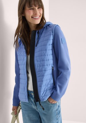 Mujer sonriente con una chaqueta acolchada azul claro con capucha, camisa blanca y vaqueros azules, sosteniendo un bolso beige con una mano.