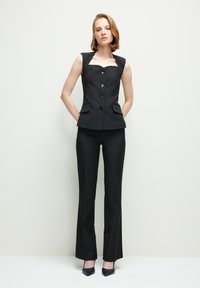 Zwarte mouwloze top met een vierkante halslijn, vijf knopen en twee voorzakken, gecombineerd met zwarte op maat gemaakte trousers en puntige hakken.