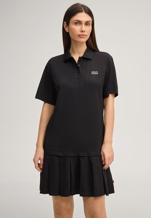 Frau mit glattem dunklem Haar, die ein schwarzes Polokleid mit kurzen Ärmeln, plissiertem Saum und "JOOP! JEANS"-Logo auf der Brust trägt.