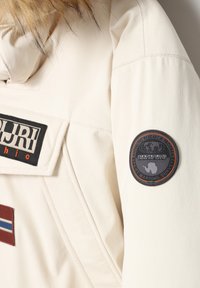 Hvid jakke med en faux fur hætte, der har sorte og røde lapper, struktureret stof og et cirkulært logo på ærmet.