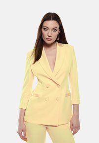 Blazer giallo doppiopetto con scollatura a V profonda, maniche lunghe e due tasche frontali. Texture liscia e design aderente.