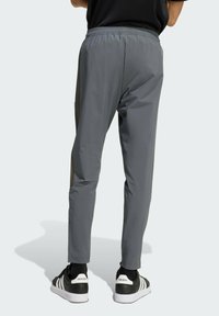 Osoba v sivých elastických nohaviciach s pásom a čiernych teniskách Adidas s bielymi pruhmi, stojaca na svetlom pozadí.