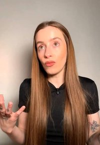 Junge Frau mit langen braunen Haaren gestikuliert mit den Händen, schaut nach oben, trägt ein schwarzes Top, vor einem schlichten hellen Hintergrund.