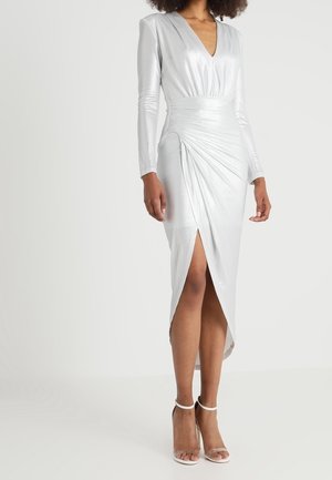 Mujer con un vestido plateado de manga larga con escote en V profundo y abertura frontal, combinado con sandalias blancas de tacón alto y tiras en el tobillo.