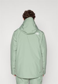 Chaqueta impermeable de color verde claro con capucha, que presenta un logo en la parte posterior y un corte recto. La tela parece suave con un ligero brillo.