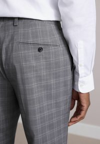 Pantalons gris à carreaux avec une texture lisse, dotés d'une poche arrière avec un bouton noir. Manche de chemise blanche partiellement visible avec des boutons.