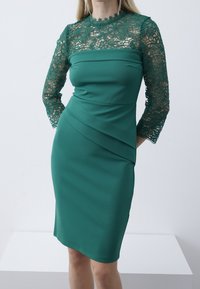 Robe verte ajustée avec manches en dentelle trois-quarts, motif floral et texture lisse ; design drapé qui met en valeur la silhouette.
