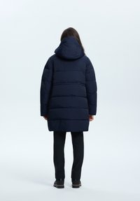 Lång marinblå pufferjacka med huva, med en quiltad design och tjock stoppning. Bärs över svarta byxor och ankelstövlar.