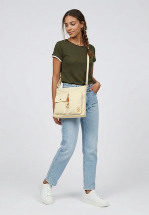 Kvinde med flettet hår iført grøn t-shirt, lyseblå jeans, hvide sneakers, bærer beige crossbody taske med forlommer og står på hvid baggrund.