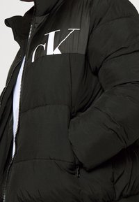 Sort dunjakke med høj krave, med en skinnende øverste del og en fremtrædende hvid "CK"-logo på brystet. Lynlåslommer inkluderet.
