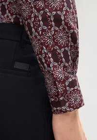Bordeauxfarbendes Hemd mit floralem Muster und langen Ärmeln sowie Knopfmanschetten, kombiniert mit schwarzen Hosen. Verfügt über zarte weiße Blumen- und Blätterdesigns.